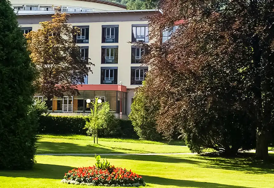 Einen Garten mit grünem Rasen, bunten Blumen und großen Bäumen. Im Vordergrund befindet sich ein Blumenkugel mit roten