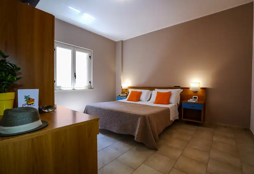 Ein Zimmer im Hotel mit einem Bett mit orangefarbenen Kissen und einem Holzbügele. Ein Schreibtisch mit einem Hut und einem