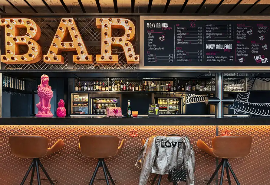 Ein Bar im Hotel Moxy Bremen mit einem leuchtenden "BAR" Lettering und einer Auswahl an Getränken und Snacks auf dem
