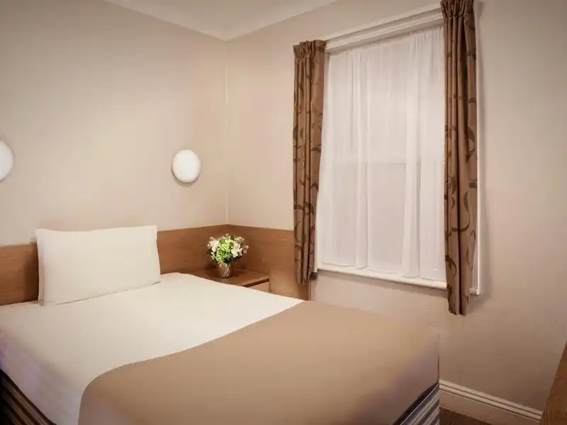 Ein Zimmer im Hotel Lords in London mit einem Bett, einem Nachttisch und einem Fenster mit Vorhängen