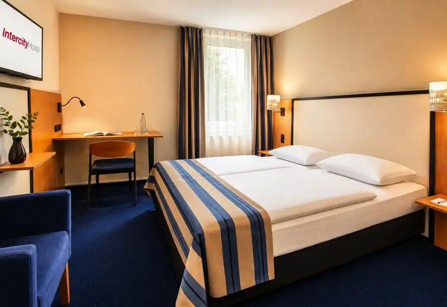 Ein Zimmer im IntercityHotel Celle. Das Zimmer verfügt über ein großes Bett mit weißem Bettlauf und blauen Streifen,