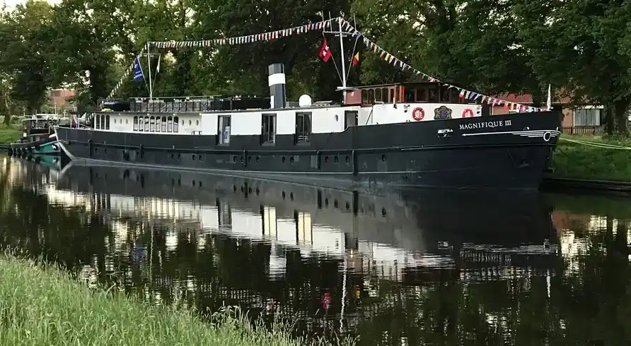 Ein großes Holzschiff namens MS Magnifique II, III oder IV an einem ruhigen Kanal in Amsterdam. Das Schiff ist schwarz und