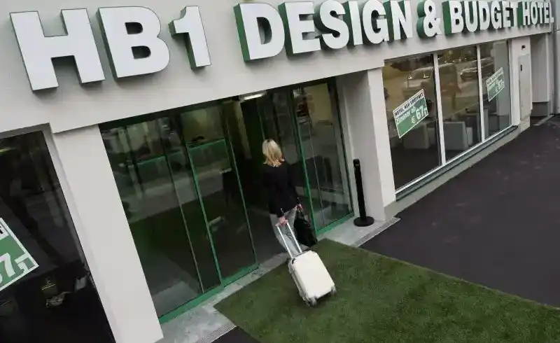 Das HB1 Design & Budget Hotel Wien-Schönbrunn. Eine Frau rolls ein Reisekoffer vor sich und steht vor den Eingängen des