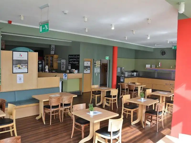 Einen Restaurantbereich mit Holenzimmern und Tischen. Der Raum ist mit hellen Farben gestrichen und mit Lichtern