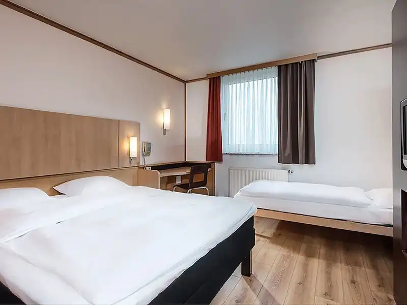 Ein Zimmer in einem Hotel. Es gibt zwei Betten, eine größere und eine kleinere, beide mit weißen Bettwäsche ausgestattet.