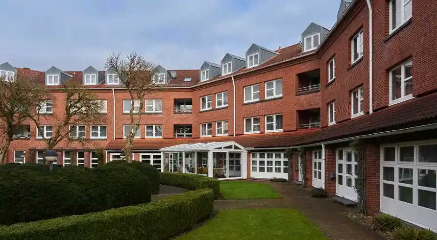 Ein großes, rotes Backsteingebäude mit mehreren Stockwerken und Balkonen. Es liegt in einem Gartenbereich mit gepflegten