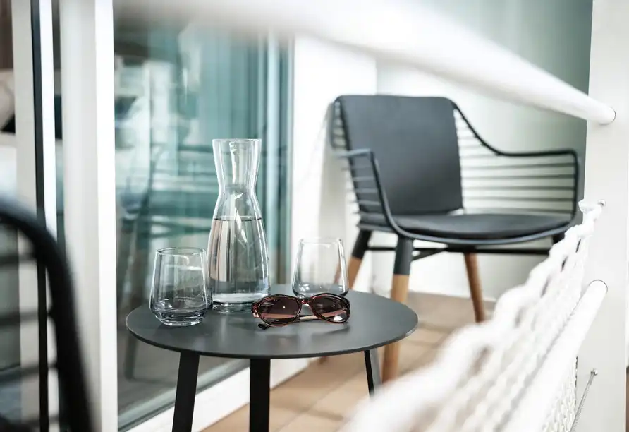 Einen Außenbereich mit einem Tisch und Stühlen. Auf dem Tisch befindet sich ein Wasserkrug, zwei Glasbecher und