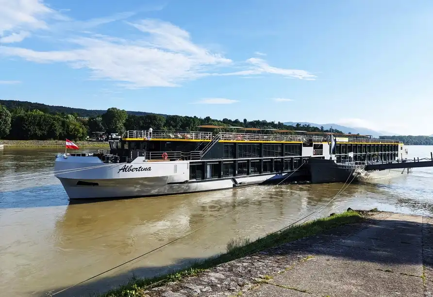 Einen großen Passagierboot, das MS Albertina genannt wird, auf der Donau. Der Boot ist teilweise an einem Ufer abgestellt,