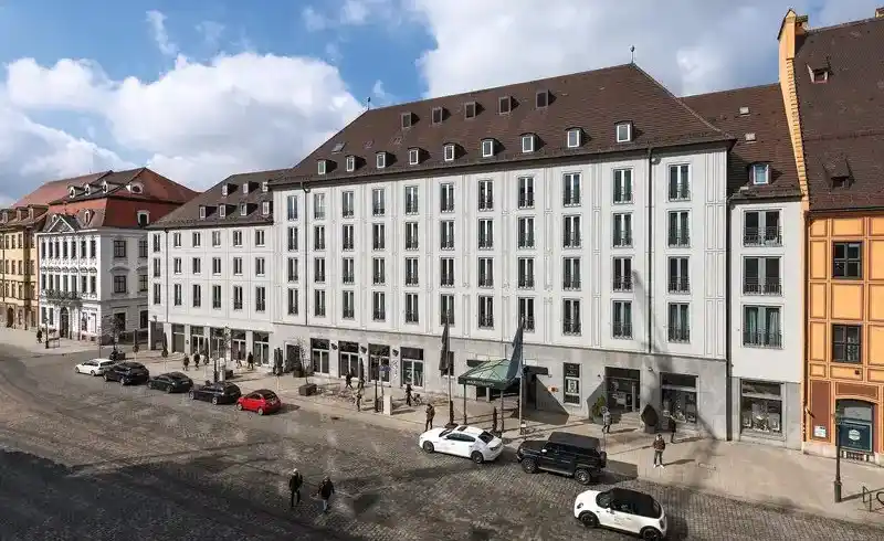 Das Hotel Maximilian's in Augsburg, einem stilvollen Gebäudewinkel mit weißgeziegelter Fassade und schwarzen Dachziegeln.