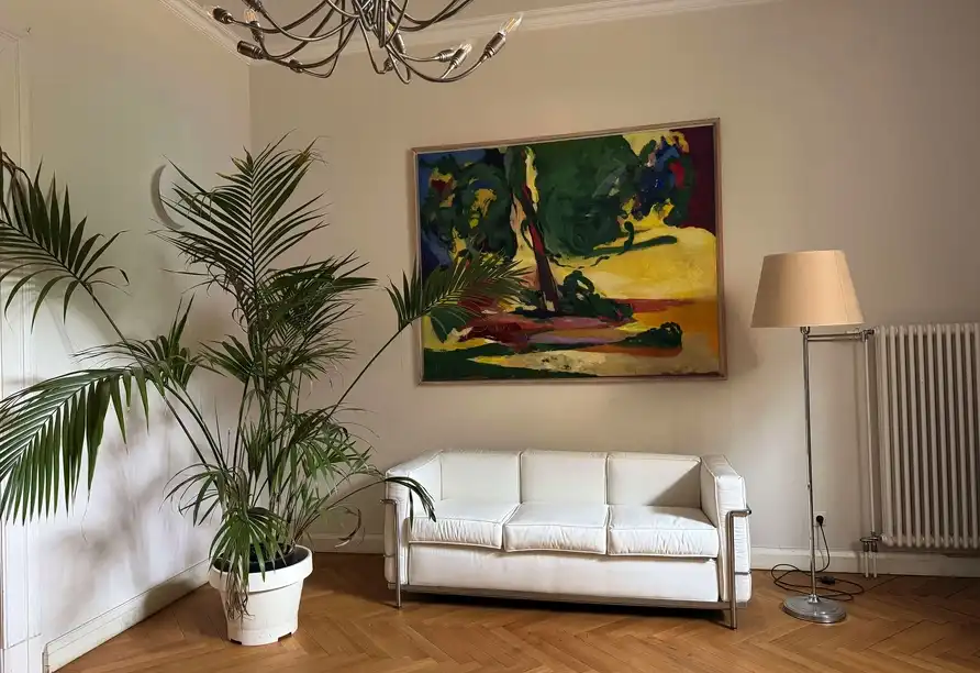 Das Zimmer zeigt ein gemütliches Ambiente mit einem weißen Sofa, einem leuchtenden Malerwerk an der Wand und einem großen