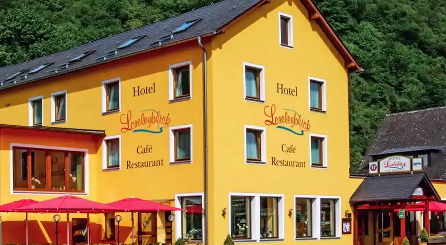 Das Hotel Loreleyblick in St. Goar, einem gelben Gebäude mit roten und rosa Sonnenschirmen vor dem Café Restaurant. Der