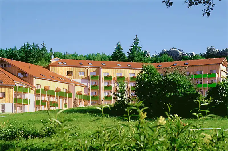 Ein großes, gelbes und orangefarbenes Sporthotel mit mehreren Gebäuden und Balkonen. Die Umgebung ist grün und natürlich,