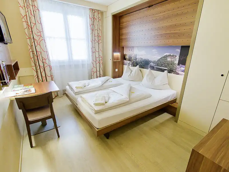 Ein Zimmer im JUFA Hotel Murau mit Blick auf ein großes Bett mit weißen Bettwäsche und einem Holzbügel. Neben dem Bett ist